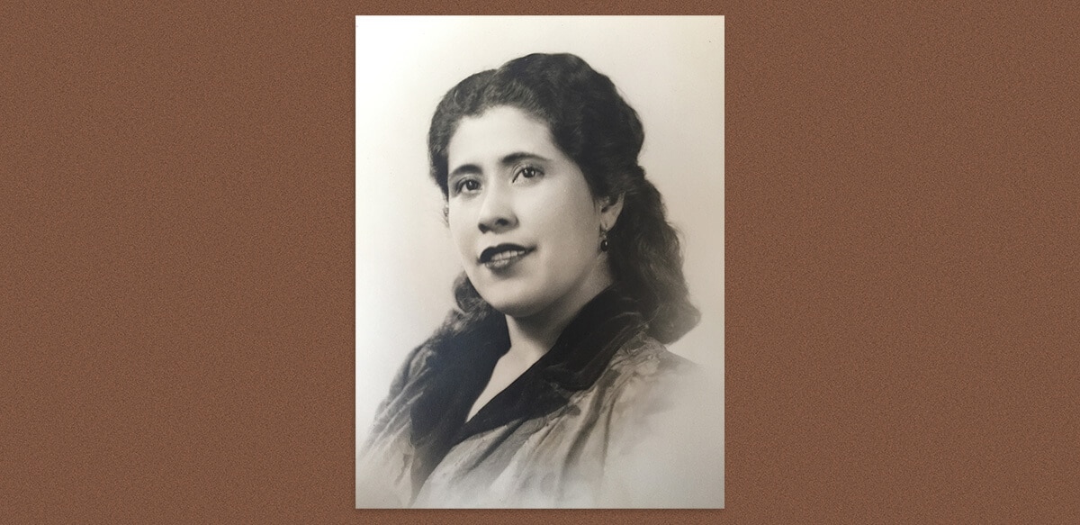 A portrait photo of a young woman with dark, wavy hair dressed up, turning to face the camera