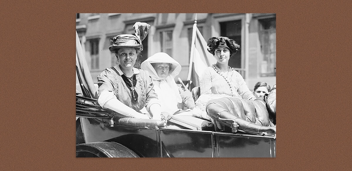 Three women in an open-topped vehicle, dressed in 1910 attire